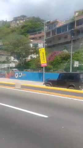 The video shows a roadside view from a moving vehicle, likely a car or bus, on a sunny day. The foreground is dominated by a paved road with white lane markings and yellow and black striped traffic barriers. In the background, there is a colorful mural on a wall, depicting abstract shapes and sporting imagery like soccer balls and basketballs. Behind the wall, there is a green grassy area with trees and some buildings, including multi-story structures situated on a hillside. Further up the hill, more houses and vegetation are visible. The video captures a brief glimpse of urban or suburban scenery with elements of recreation and residential areas.
