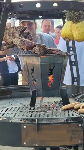 A man wearing a brown leather apron and a baseball cap smiles as he holds up a large piece of meat skewered on a metal rod over a grill. The meat appears to be cooking on a rotisserie, suspended above a fire pit with glowing coals. In the background, several people watch, some smiling. Two pineapples hang from a metal rack to the right of the main grill. Below the rotisserie, a smaller grill contains several sausages cooking over coals. The scene appears to be an outdoor event, possibly a food festival or market, with other vendors and people visible in the distance. The overall impression is of a lively, open-air culinary demonstration.
