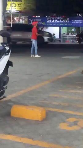 A person wearing a red t-shirt, blue jeans, and a red cap stands in a paved outdoor area. They are holding a yellow object, possibly a toy or a signaling device, and appear to be waving it or throwing it. In the background, a silver car is driving past, and there are buildings with signs, including one that says "SIJICOM" and another with Japanese characters. The ground is marked with yellow lines and a yellow concrete barrier is in the foreground. The lighting suggests it is evening or nighttime.