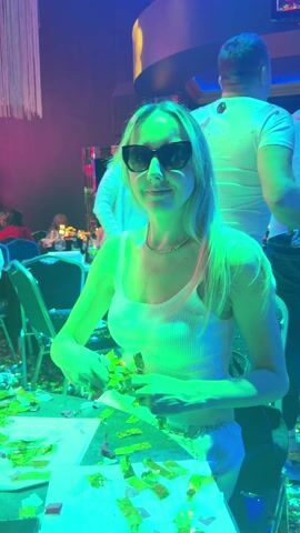 A woman with long blonde hair and wearing sunglasses, a white ribbed tank top, and white pants stands at a table in an event space, cheerfully throwing confetti into the air. The confetti is a mix of gold and green. The table is covered with confetti as well as printed materials. Other people dressed in white can be seen in the background, suggesting a themed party. The venue has dim lighting with pink and purple accents, and a decorative chandelier hangs in the background. A music track plays in the background.