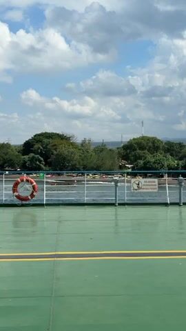 The video shows a view from the deck of a ferry or boat, looking towards land. The deck is painted green with yellow stripes. A life preserver, which is orange and white, is hanging on a railing. Beyond the railing is a body of water, and then the shoreline with trees and some buildings. The sky is blue with white clouds. There are no people visible in the video. The camera is stationary.