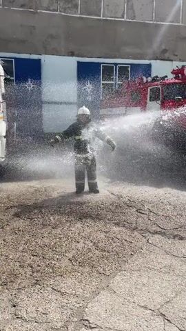 The video captures a firefighter in full gear, wearing a helmet and protective suit, standing in a cloud of water. The firefighter is being sprayed from both sides by water jets, creating a dynamic and misty effect. The activity appears to be a training exercise or demonstration, possibly involving testing equipment or practicing hose handling.

The setting is an outdoor area with a rough, gravelly surface and some cracked concrete in the foreground. In the background, two red fire trucks are partially visible, suggesting this is a fire station or training ground. The building behind them is light-colored with blue panels and windows, featuring a star emblem on the blue panels. The lighting suggests it is daytime, with bright sunlight casting shadows. The overall atmosphere is one of controlled action and perhaps a bit of playful energy, given the way the firefighter is positioned amidst the water spray. There are no visible signs with text in the frame. The scene is likely in Chusovoy, Russia, as indicated by the location context.