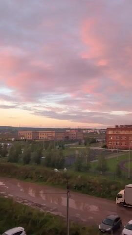 The video displays a wide shot of a suburban landscape at dusk. The sky is filled with a gradient of pink and purple clouds, indicating the setting sun. In the foreground, a muddy road with puddles is visible, with a few cars parked nearby. Beyond the road, there is a grassy area with trees and a sports field, which appears to be a soccer field. Several buildings, possibly schools or apartment complexes, are visible in the midground. A white truck is parked on the right side of the road. The overall scene evokes a peaceful, serene atmosphere.