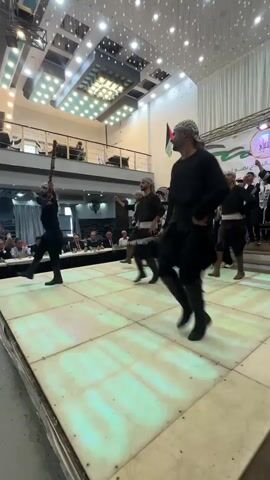 The video shows a group of men dancing on a brightly lit stage. The dancers are all wearing black clothing, including long tunics, pants, and tall boots. Many of them are holding long, thin objects, possibly sticks or rifles, which they are moving in sync with their dance. The stage is white and appears to be made of illuminated panels. In the background, there is an audience seated at tables, and further back, a balcony with more people. The venue appears to be a hall or ballroom, with decorative lighting on the ceiling and a large banner with Arabic text and a logo hanging on the back wall. A Palestinian flag is also visible. The overall atmosphere seems to be festive and celebratory.