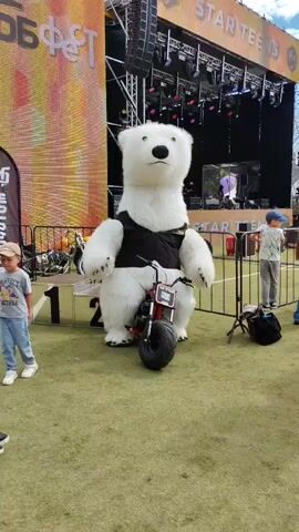 A person in a polar bear mascot costume stands next to a small red motorcycle. A person in a wheelchair approaches the mascot. The mascot turns to face the person in the wheelchair, who then rides past the mascot. A child in the background dances. The scene is outdoors, in front of a stage setup.