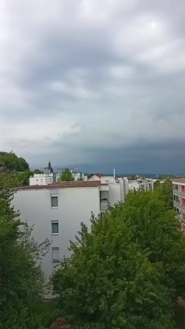 This media file shows an aerial view of a residential area in Zofingen, Switzerland. The scene is dominated by overcast skies with dark storm clouds gathering on the horizon. In the foreground, several apartment buildings are visible, characterized by their white facades and balconies with pink railings. Lush green trees are abundant, obscuring the lower floors of some buildings and contributing to the overall natural setting within the urban environment.

The activity depicted is primarily static, focusing on the urban landscape and the impending weather event. The time of day appears to be daytime, given the ambient light, but the heavy cloud cover creates a dim, somber atmosphere. The weather is clearly stormy or about to become so, with the dark clouds suggesting rain or a thunderstorm.

Notable details include the architecture of the apartment buildings, with their repetitive balcony designs and flat roofs. The presence of trees indicates a well-maintained green space within the town. In the distance, the green hills of Zofingen are visible, dotted with houses. A few faint plumes of smoke or steam rise from industrial or residential areas in the far distance, indicating some level of human activity beyond the immediate residential blocks. The overall mood evoked by the dark clouds and the overcast sky is one of anticipation and perhaps a slight sense of foreboding. There is no visible text in the media file.