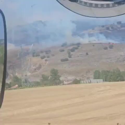The video opens with a reflection of a vehicle's interior and mirrors, showing a road. The scene then shifts to an outdoor view of a dry, hilly landscape. Smoke is visible rising from a distant hillside, indicating a fire. Power lines and pylons are present on the landscape. In the foreground, a harvested field of dry grass or wheat is visible. A few buildings, possibly houses, are scattered in the middle ground, surrounded by trees. The overall impression is of a rural area experiencing a wildfire.