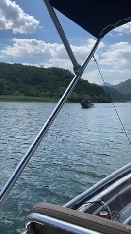 This media file shows a scenic view of a body of water, likely a river or lake, on a sunny day with scattered clouds. The perspective is from a boat, indicated by the presence of a metal railing and a portion of the boat's canopy in the foreground.

The water is a deep blue with ripples, suggesting a gentle breeze. In the distance, a lush green, tree-covered hill forms the backdrop. The vegetation appears dense and vibrant, indicative of a warm season. On the left side of the frame, another boat can be seen with several people on board, creating a white wake as it moves across the water. The scene is peaceful and suggests recreational activity on the water.

The time of day appears to be daytime, likely late morning or afternoon, given the brightness of the sun and the shadows. The weather is pleasant and fair, with the clouds adding visual interest to the sky without obscuring the sunlight. There are no visible text elements in this media file. The overall mood is one of relaxation and enjoyment of nature and outdoor activities. The location context provided is Riječani, Montenegro, which aligns with the natural landscape depicted.