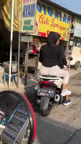 The video shows a person, likely female based on the clothing, wearing a hijab and a black top and white pants, sitting on a white scooter. The scooter has a license plate that reads "W 5797 MDO". The person is facing away from the camera, looking towards a food stall. The food stall has a yellow awning with text that appears to be in Indonesian, possibly advertising "AYAM" (chicken). There are menus and other items displayed at the stall. The scooter's rear is in the foreground. The scene appears to be outdoors, possibly on a street or in a market area, with a sunny sky. A reflection of another scooter's mirror is visible in the bottom left corner of the frame, showing a reflection with text and images. The overall atmosphere is casual and daytime.