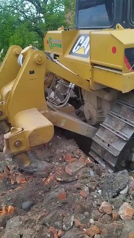 A yellow Caterpillar bulldozer, model D6R2, is partially submerged in muddy water and debris. The bulldozer is angled, with its tracks sunk into the muck. The front blade appears to be resting on a piece of concrete or rubble, lifting the front of the machine slightly. The tracks are caked with mud. Debris, including broken bricks and dirt, is scattered around the bulldozer. The surrounding environment appears to be an outdoor construction or demolition site, with hints of greenery in the background. The video captures close-ups of the bulldozer's tracks and undercarriage, emphasizing its stuck position. No people are visible in the footage.