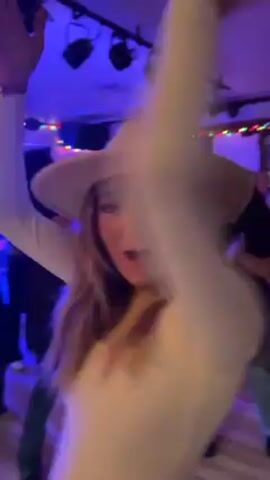 A woman with long blonde hair and a beige cowboy hat is seen dancing at a party. She wears a long-sleeved white top. The party appears to be indoors with multiple people around. A man with a beard wearing a black cap moves through the crowd. The lighting is dark, with blue uplighting and string lights visible. Other people are dancing in the background.