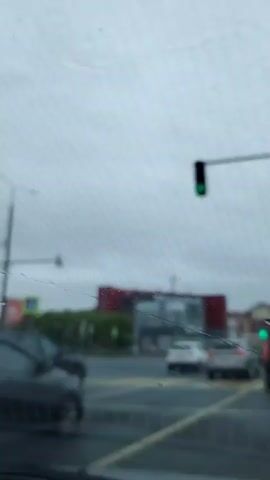 The video appears to be shot from inside a vehicle, looking through a rain-streaked windshield. The weather is overcast and appears to be raining. The scene shows a blurred intersection with several cars. A traffic light is visible, showing a green signal. Buildings and trees are visible in the background, also blurred. The focus is on the windshield and the conditions outside, rather than a clear view of the surroundings.