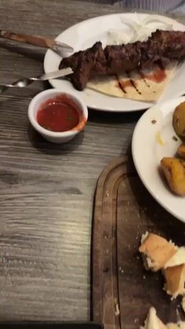 The video shows a wooden table with a meal laid out on it. There is a plate of meat kebabs on a skewer, served on flatbread with sliced onions, and a small dish of red sauce. Another plate holds chunks of roasted potatoes with herbs.  Bread slices and a small container of butter sit on a separate plate.  A person's hand holding silverware is visible at the upper left.