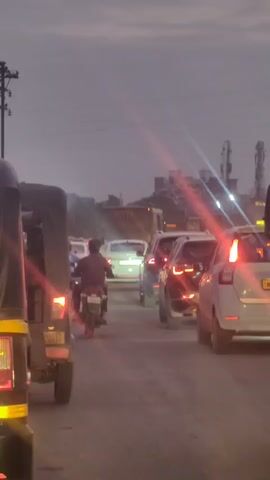 The video shows a street scene during twilight. There is moderate traffic including cars, motorcycles, and an auto-rickshaw. The sky is overcast with a purplish hue, suggesting the end of the day. Streetlights and vehicle headlights are illuminated, creating lens flares in the video. The traffic appears to be moving slowly or is stopped, implying congestion. The overall environment suggests an urban or semi-urban setting with some buildings and utility poles visible in the background.