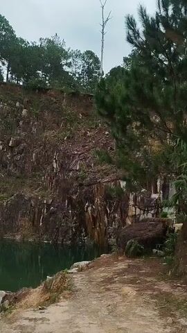 A person wearing a yellow hat, blue shirt, and khaki shorts with a backpack stands on the edge of a quarry, holding a stick. The quarry has steep, rocky walls and a body of green water at the bottom. Trees grow along the top of the quarry rim. The camera pans across the quarry, showing more of the rocky landscape and the water. There is a small raft floating on the water in the distance.