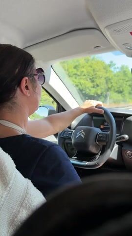 A person with dark hair and sunglasses is driving a car. They are wearing a dark blue shirt and have a white strap around their neck. The steering wheel is black with a Citroen logo. The dashboard shows a digital display. Outside the car, trees and a blue sky are visible, along with another car in the distance. The interior of the car is visible, including a white towel or blanket on the left side. The lighting suggests it is daytime.