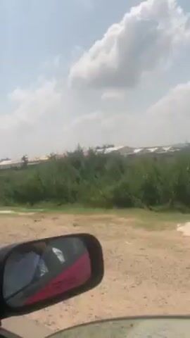 The video, taken from the inside of a car, shows a view from the passenger seat looking out the side window and forward. The car is driving on a dirt road, and the landscape features dry, grassy terrain with some green vegetation. In the distance, a row of buildings is visible under a partly cloudy sky. The car's side mirror is visible in the foreground, reflecting the interior of the car and a person wearing a white shirt. The camera is handheld, resulting in some movement as the car drives.