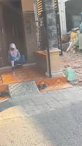 A young girl wearing a pink hijab and a blue and white school uniform sits on the steps of a house. She is looking down at something in her lap. A black bag is on the ground next to her. The house has a brick facade and a tiled roof. There are several pairs of sandals lined up on the steps. The area around the house appears to be under construction or renovation, with piles of rubble and building materials visible. The sun is shining, casting long shadows.