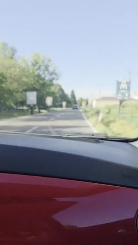 The video shows a first-person view from inside a car, looking forward through the windshield. The road ahead is asphalt with white lane markings. Trees and foliage line both sides of the road, and some buildings are visible in the distance on the right. A few other cars are on the road ahead, and one car is passing in the opposite direction. There are signs visible along the road, including a large billboard on the left and smaller signs on the right. The dashboard of the car, which is red, is visible in the foreground. The sky is clear and blue, indicating a sunny day.