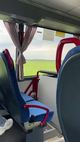 From inside a bus, the camera focuses on the seat directly in front, capturing a view out the window. The window reveals a green field stretching into the distance, dotted with occasional structures. The initial focus is on the bus interior, with a blue and red seat in the foreground, partially obscuring the view. As the bus moves, the camera’s perspective shifts to center the outdoor scenery. The green field is bordered by a wire fence, and beyond that, there are a few scattered houses and some trees in the background. The sky is overcast.