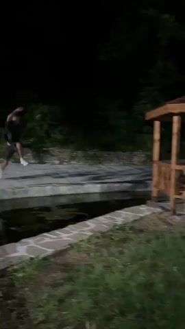 A person is seen jumping into water from a ledge. The person lands in the water and then emerges. The person is wearing a grey shirt and black shorts with white shoes. The person then walks away from the water and sits down on the grass. The scene takes place at night and is lit by artificial lights. There is a wooden gazebo in the background.