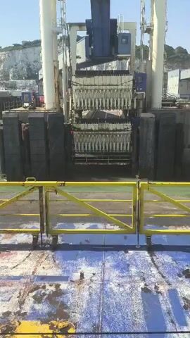 A ferry or large ship's deck is in the foreground, with peeling blue and white paint and a yellow safety railing. Beyond the railing is a dock with large, dark grey blocks forming a quay. A large white structure with blue accents, resembling a gantry or crane, is situated on the dock. This structure has multiple rows of what appear to be hydraulic arms or pistons. The sea is visible behind the structure, and a cliff face, likely white chalk, is in the background on the left. The sky is clear and blue. The number "9" is visible on the white gantry.