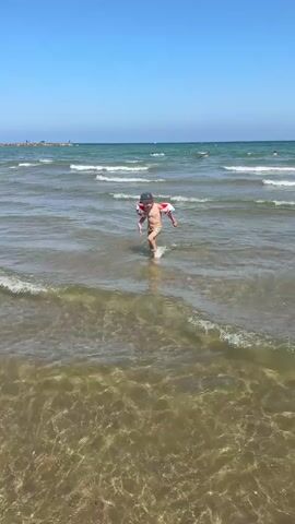 A young boy wearing a dark hat and swim trunks with a cartoon pattern runs into the shallow ocean water. He has inflatable armbands with a Mickey Mouse design on them. The sun is shining brightly in a clear blue sky, indicating it is daytime, likely midday or early afternoon. Gentle waves are rolling in, and the water is clear enough to see the sandy bottom. In the background, a breakwater or pier stretches out into the sea, with some people visible in the distance. The overall scene suggests a pleasant day at the beach, with the boy appearing happy and engaged in his activity. The location is identified as Urbanització Port Saplatja, Spain. There is no visible text in the media.