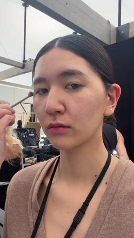 Here is a description of the video:
A young Asian woman is the focus of the video, standing in a backstage area. She has dark hair pulled back, fair skin with some blemishes, and is wearing a light brown cardigan with a black lanyard around her neck.  She looks directly at the camera, and then smiles slightly.

Around her, several people are working. To her left, a fair-haired woman with blonde hair is seated and appears to be getting ready. Behind her, a man with dark hair is applying makeup to another woman. Another pair of hands holding a makeup brush are just to the left of the woman's face, close to her left eyebrow.

The background shows a busy scene, with makeup kits, mirrors with lights, and other equipment typical of a backstage or dressing room environment. The lighting is bright, and the overall atmosphere suggests preparation for a fashion show or photoshoot.