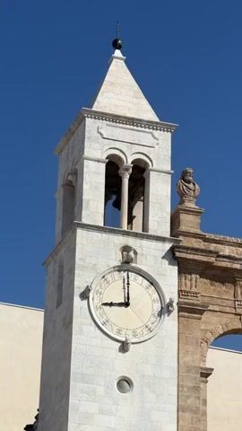 A video shows a close-up view of an old clock tower against a clear blue sky. The clock has Roman numerals and the hands indicate approximately 9:08. To the right of the clock tower, a section of an ancient stone archway is visible, adorned with two stone busts. The video pans slightly upwards, revealing more of the clear blue sky. The overall scene depicts a historical architectural feature in bright daylight.
