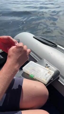 The video shows a person sitting in an inflatable boat on the water. The person is wearing a life vest and a blue and white cap. They are using their hands to tie something to a fishing line. Next to them is a tackle box with fishing lures inside. The water is slightly choppy and the sun is shining. The person's arms and legs are visible, and they are wearing dark shorts.