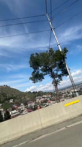 The video shows a landscape from a moving vehicle, likely a car. A concrete barrier runs along the bottom of the frame. The view opens up to a cityscape with buildings of varying heights and colors. A small, green hill with sparse vegetation is on the left side of the frame. The sky is blue with scattered white clouds. The wind is strong, as indicated by the audible wind noise. There are trees scattered throughout the cityscape and along the hillside. Power lines can be seen in the sky.