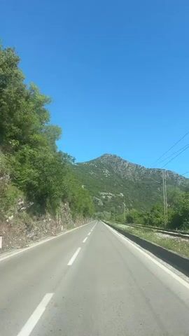 The video shows a view from a car driving on a straight, paved road towards a mountain under a clear blue sky. On the left side of the road is a rocky embankment with green trees and vegetation. A white line marks the center of the road. On the right side of the road is a metal guardrail and more vegetation. Power lines are visible on the right side of the road. The mountain is covered in green vegetation and rocky outcroppings.