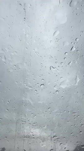 This is a close-up, vertical video of raindrops on a window pane. The raindrops are of various sizes, from small droplets to larger, elongated streaks. The background is blurry and appears to be a cloudy sky or an overcast environment. The lighting is dim and diffused, consistent with a rainy day. The video shows the natural movement of the raindrops, with some appearing to be sliding down the glass and others remaining static. There are no people or other objects visible in the frame. The overall impression is one of a rainy, somber atmosphere.