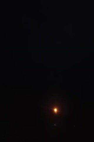 The video opens to a dark sky with a single, bright orange light in the center. This light appears to be a streetlamp or a similar artificial light source. The camera then pans downwards, revealing a silhouetted roofline of a building against a hazy, illuminated sky. Trees are also visible as dark shapes against the light. Another bright orange light, consistent with the one seen earlier, is visible near the roofline. The video then returns to the dark sky with the single orange light.