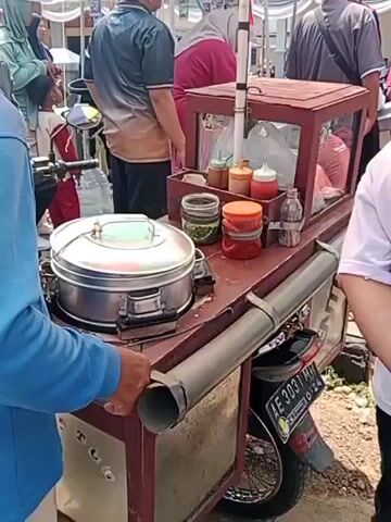 The video shows a food cart parked outdoors, possibly at a market or street event, with several people around it. The cart is mounted on a motorcycle and appears to be selling steamed food, with a large metal steamer prominently displayed. On the cart's surface, there are various containers of condiments, including jars of chopped green chilies and what appear to be sauces or relishes. A glass display case sits on the cart, containing more food items and utensils. The people in the video are dressed casually, and some seem to be interacting with the food vendor or browsing the offerings. The environment is bright, suggesting daytime, with some tents or awnings visible in the background. The license plate of the motorcycle is visible, indicating it is an Indonesian vehicle. The overall scene depicts a common street food vending operation.