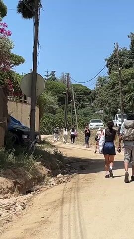 A group of people are walking down a dirt road. The road is lined with trees and plants. There are utility poles with wires overhead. The sky is clear and blue. A black car is parked on the left side of the road.