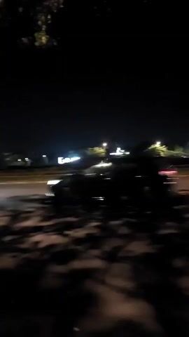 A night-time video shows a car driving on a road. The car appears to be black and is moving from left to right in the frame. The road is paved and there are streetlights visible in the background. There are trees on the left side of the road, and their shadows are cast on the road, creating a dappled effect. The sky is dark and there are some lights visible in the distance, possibly from buildings. The video is shaky, suggesting it was recorded while in motion.