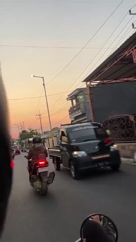 The video shows a street scene at sunset. In the foreground, the perspective is from someone on a motorcycle. To the left, a person on a scooter is riding away from the camera. To the right, a black pickup truck is driving past. In the background, there are buildings, streetlights, and power lines. The sky is a soft orange and pink, indicating sunset. The road appears to be paved and is wet in places. The overall impression is that of a typical urban or suburban street during the golden hour.