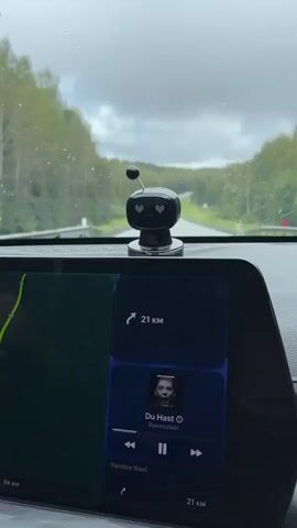 The video begins with a close-up view of a car's dashboard. A small, black, robot-shaped air freshener with heart-shaped eyes sits on the dashboard, facing forward. The car's windshield wipers are active, and raindrops are visible on the windshield. In the background, a winding road stretches through a forest under a cloudy sky. The car's infotainment screen displays navigation information, showing a route with "21 KM" remaining, and a music player interface showing the song "Du Hast" by Rammstein. The camera is positioned inside the car, looking out through the windshield. The overall scene depicts driving in rainy weather.
