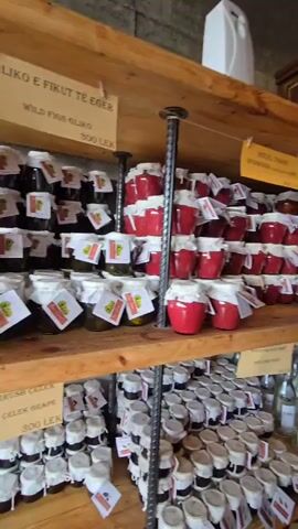 Here's a concise description of the video:

The video shows the interior of a shop selling locally-made goods.  Shelves are stacked with jars of jam, preserves, and bottles of wine or syrup.  People are browsing the shelves and standing around crates of fresh cherries and strawberries.  The shop has a rustic feel with exposed concrete ceilings and wooden shelves.  One woman is examining the shelves closely.