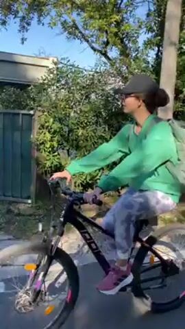 A young woman with a baseball cap, green long-sleeved shirt, and tie-dye pants is riding a bicycle down a paved road. She's wearing a backpack and has pink shoes on. To her right is a building with "RECEPTION" written on it, along with a sign that reads "AUTO CAMP DULE". In the background, a mountain range is visible under a clear blue sky. The camera moves slightly, following the woman as she continues to ride.
