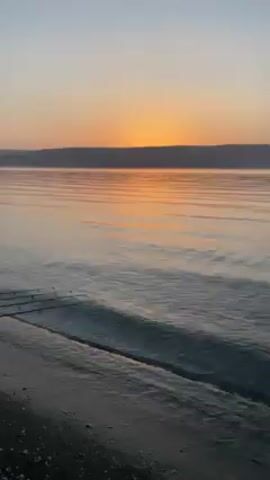 The video captures a serene scene of a body of water, likely a lake or sea, at sunrise. The sky transitions from a pale blue to a warm orange hue as the sun begins to emerge behind a dark, distant landmass. The water's surface is calm with gentle ripples reflecting the colors of the sky. The shoreline consists of a dark, sandy beach. Several fishing rods are visible in the foreground, resting near the water's edge, suggesting someone is engaged in fishing. The overall atmosphere is peaceful and tranquil.