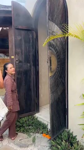 A person opens a large, ornate wooden door to reveal a passage leading to a small, circular pool surrounded by lush greenery. The passage is tiled and has a plain white door at the end. The pool is partially covered by a thatched roof structure, and decorative orange flowers are placed on the stone border.