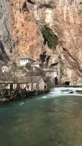 The video shows a fast-flowing river in front of a rocky cliff face. Several buildings are nestled against the base of the cliff, and the river appears to flow out from under the cliff near one of the buildings. The buildings are constructed of stone and appear to be old. The river is a clear turquoise color. The cliff face is a mix of light and dark brown rock, with some greenery growing on it. The water is churning and splashing as it moves over the rocks in the riverbed. The weather appears to be sunny.