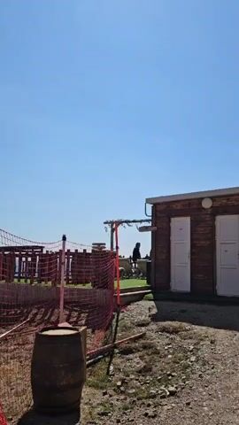 A person is seen from behind, sitting at a table inside a small, wooden building with large windows. The building appears to be a restroom or service facility. Outside, a ski lift tower and chairs are visible, suggesting a mountainous or ski resort environment. The sky is clear and blue, indicating fair weather. In the foreground, a barrel and some fencing are present, along with a gravelly, rocky ground. The camera is panning from left to right, showing more of the ski lift structure as it moves.