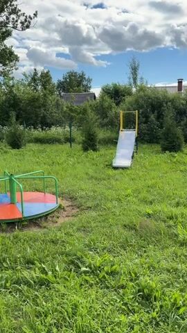 The video pans across an outdoor playground on a sunny day with a cloudy sky. In the foreground, a patch of green grass is visible, with some patches of weeds growing through. To the left, a red and green children's spinning toy is partially visible. Further back, a yellow and silver slide stands in the grass, with a swing set to its right. A red and green caterpillar-shaped bench is in the middle ground. The camera then moves to show a tiled path with weeds growing between the tiles, leading towards a sports court. On the court, a soccer goal and basketball hoop are visible. Two children are briefly seen on the court. The background consists of trees and houses.