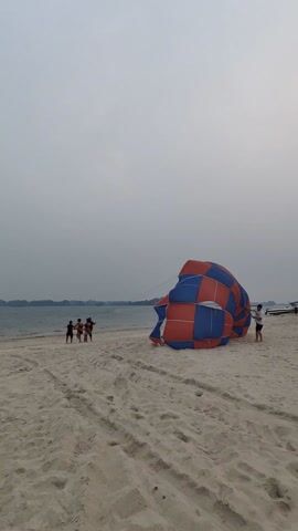 The video shows a beach scene with a red, blue, and white parachute being filled with air. The parachute has a checkered pattern. On the sand, three people are holding onto ropes attached to the parachute, seemingly trying to keep it steady. A man in a white shirt is standing nearby, holding a pole. In the background, there's a body of water with visible land in the distance and a boat is near the right edge of the frame. The sky is overcast. There are multiple tire tracks in the sand. Another person can be seen taking a photo in the distance, on the left.