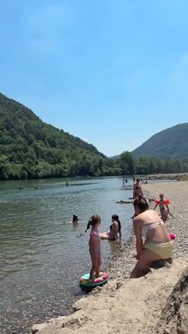 This media file captures a sunny day at a river beach, likely in Zvornik, Bosnia and Herzegovina, given the context. The scene is vibrant with activity, as several people are enjoying the water and the shoreline on a clear day.

The primary subjects are people, including adults and children, engaged in various recreational activities. In the foreground, a woman in a yellow bikini is crouching on the rocky shore, facing away from the camera. Beside her, a young girl in a pink swimsuit stands on a colorful inflatable toy in the shallow water. Other individuals are scattered throughout the river and along the beach. Some are swimming, while others are wading or relaxing by the water's edge. A few children are visible in the water, one with red floatation devices on their arms.

The setting is a wide river flanked by lush, green, tree-covered hills. The water appears calm and inviting, reflecting the bright blue sky. The shoreline is a mix of sand and smooth pebbles, typical of a river beach. In the background, more people can be seen further up the river, suggesting a popular spot for recreation.

The overall atmosphere is one of leisure and enjoyment. The bright sunlight and clear skies indicate pleasant weather, perfect for a day outdoors. The presence of swimwear, floatation devices, and the relaxed postures of the people suggest a casual and fun outing. There is no visible text within the media file.