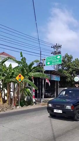 The video shows a view from a moving vehicle. The road ahead is visible with a black pickup truck in the foreground on the left. A green road sign indicates directions to Purwokerto, Yogyakarta, Cilacap, and Bandung. There are banana trees and power lines on the left side of the road, with buildings and shops on the right. Indonesian flags are displayed on poles. A white car and a yellow truck are seen driving on the road in the distance. The sky is clear and blue with some clouds. The video pans slightly to the right as the vehicle continues to move.