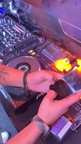 The video shows a close-up, overhead view of a DJ setup. The DJ's hands are visible, one holding a smartphone and the other resting on a DJ controller. The DJ equipment includes multiple turntables (CDJs) and a mixer, all illuminated by colorful LED lights. The background is dark and appears to be a dimly lit club or event space. The DJ is actively engaged with the equipment, suggesting they are performing or preparing to perform. The overall atmosphere is lively and energetic, typical of a DJ set.